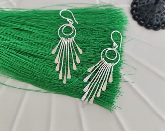 May include: A pair of silver earrings with a unique design. Each earring features a circular top with a series of dangling, elongated teardrop shapes. The earrings are displayed on a bright green textured surface.