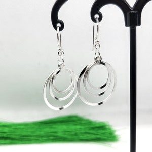 May include: Silver drop earrings with three concentric oval hoops. The earrings hang from a simple hook. The image shows the earrings against a white backdrop with a green accent.