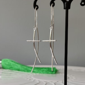 Op de afbeelding: Een paar zilveren oorhangers met een kruisontwerp. Het kruis wordt gevormd door twee gebogen lijnen die elkaar in het midden kruisen. De oorhangers hangen aan een delicate ketting.
