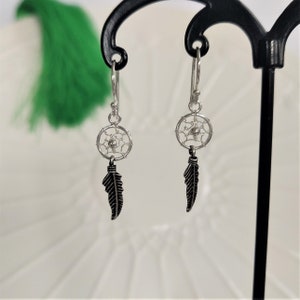 Puede incluir: Pendientes de plata con forma de atrapasueños con abalorios de plumas negras. Los pendientes están colgados de un soporte para pendientes negro.