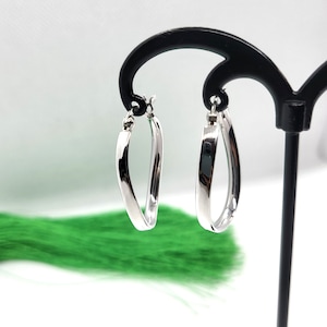May include: A pair of silver hoop earrings with a slightly oval shape. The earrings are hanging on a black earring display stand.