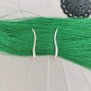 Pode incluir: Um par de brincos de prata com um design curvo ligeiramente ondulado. Os brincos estão sobre um fundo verde.