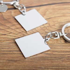 May include: Two silver-colored square keychains with a polished finish are displayed on a wooden surface. Each keychain has a square pendant attached to a chain and a keyring. The keychains are designed for personalization.