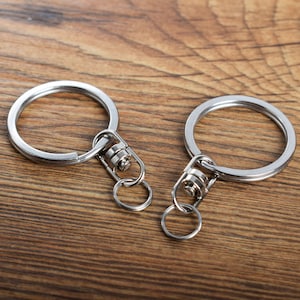 May include: Two silver keyrings with split rings and swivel clasps. The key rings are circular and made of metal. The clasps have a small ring attached for keys. The background is a wooden surface.