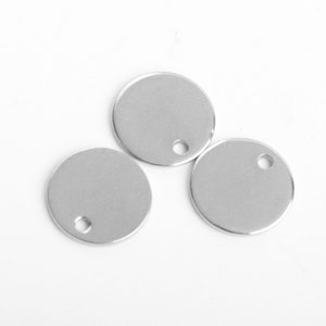 Puede incluir: Tres colgantes redondos de metal plateado brillante con un pequeño agujero perforado cerca del borde superior. Los colgantes están en blanco y listos para personalizar. Se muestran sobre un fondo blanco.