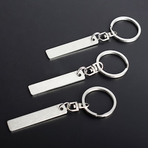 May include: Three silver keychains with rectangular plates and round key rings. The keychains are made of a shiny, brushed metal and are displayed against a dark background. The key rings are attached to the plates with small metal clasps.