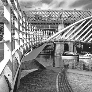 May include: A black and white photo of a bridge over a canal. The bridge is made of metal and has a curved design. There are two boats in the water below the bridge.