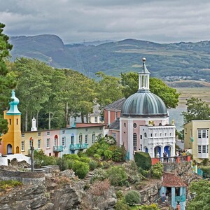 Könnte beinhalten: Ein farbenfrohes Dorf mit einem weißen Kuppelgebäude mit grünem Dach und einem gelben Turm mit blauer Spitze. Das Dorf ist von grünen Bäumen und Hügeln im Hintergrund umgeben.