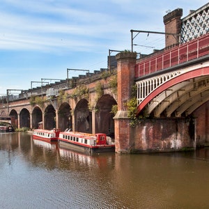 May include: A panoramic view of a canal with a red and white bridge and brick arches. Several red and white canal boats are docked in the water. The sky is blue with some clouds.