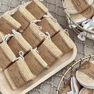 May include: A close-up of a variety of natural loofah facial pads in two baskets. The loofah pads are packaged in white muslin bags with a brown paper label that reads "LOOFAH FACIAL PADS" and includes the company's website address. The loofah pads are arranged in a variety of ways, some are stacked on top of each other, while others are spread out.