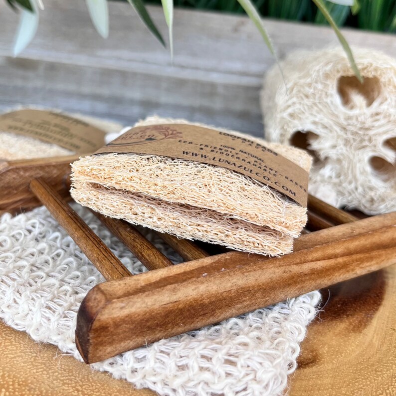 May include: Two natural loofah sponges stacked on top of each other. The sponges are light brown and have a rough texture. The sponges are sitting on a wooden soap dish. The soap dish is brown and has a rectangular shape. The text on the label reads "Loofah Sponges 100% Natural Eco-Friendly Plant-Based Biodegradable www.lunazulco.com".