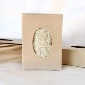 May include: A natural loofah sponge is displayed in a rectangular cardboard box with an oval cutout. The box is light brown, and the loofah is visible through the opening. Other boxes are stacked nearby, suggesting a set or collection.