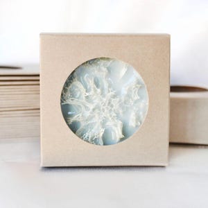 May include: A square, light brown box with a circular window revealing a light blue, snowflake-shaped object. The snowflake has white, intricate details. Other boxes are visible in the background, suggesting a set or collection.