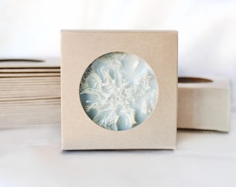 May include: A square, light brown box with a circular window revealing a light blue, snowflake-shaped object. The snowflake has white, intricate details. Other boxes are visible in the background, suggesting a set or collection.