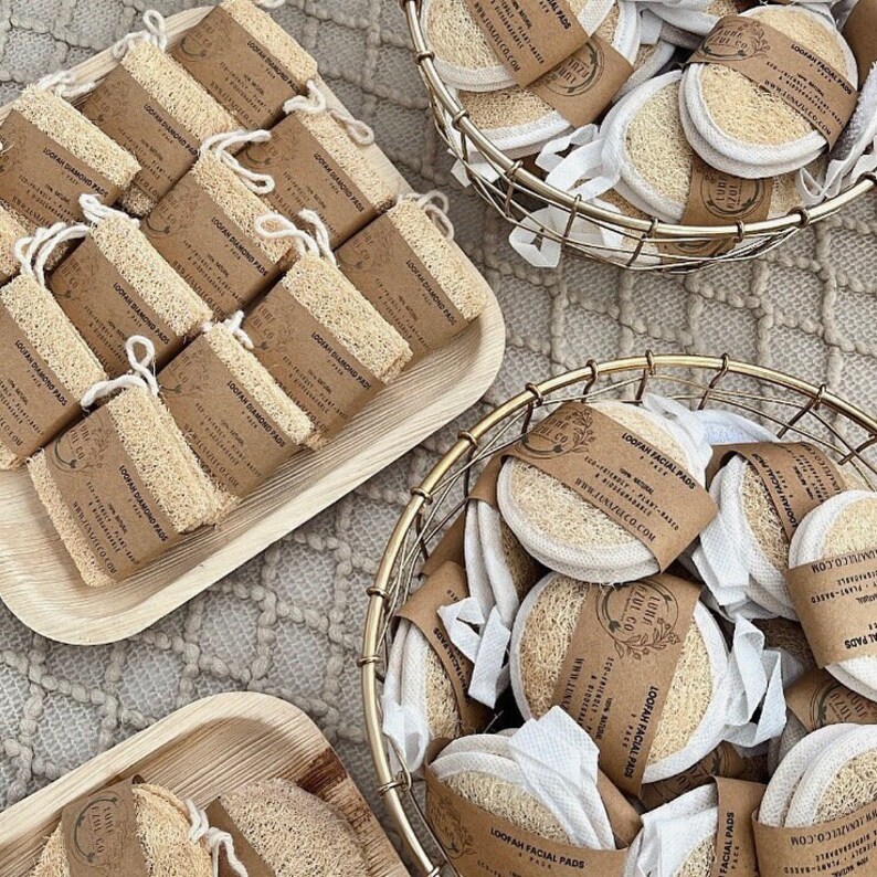 May include: A close-up of a variety of natural loofah facial pads in two baskets. The loofah pads are packaged in white muslin bags with a brown paper label that reads "LOOFAH FACIAL PADS" and includes the company's website address. The loofah pads are arranged in a variety of ways, some are stacked on top of each other, while others are spread out.