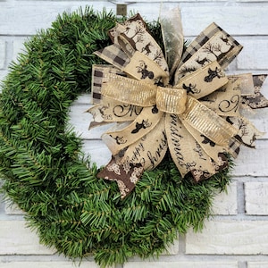 May include: A Christmas wreath made of green faux pine needles, adorned with a large bow. The bow features a gold and brown color scheme with reindeer and holiday text. The wreath is displayed against a white brick wall.