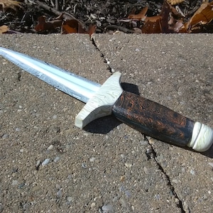 May include: A handcrafted dagger with a patterned steel blade and a wooden handle. The handle features a dark brown and orange swirl design, with white accents at the guard and pommel. The dagger is displayed on a concrete surface.