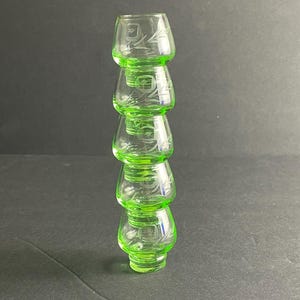 May include: A stack of four green glass shot glasses. Each glass has a rounded base and a flared top. Delicate etched floral designs adorn the sides. The glasses are arranged vertically against a dark background.