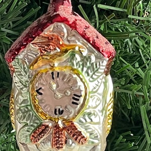 May include: A vintage Christmas ornament shaped like a cuckoo clock. The ornament is silver with gold accents and a red roof. The clock face has black hands and numerals. The ornament hangs from a red string and is surrounded by green pine needles.