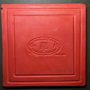 Może przedstawiać: Kwadratowa, czerwona płytka ceramiczna z zagłębioną obw&oacute;dką. Na środku owalny emblemat z napisem "Wom and Richard" i "Mackenzie-Childs Ltd.". Płytka oznaczona "PM-G 1992".