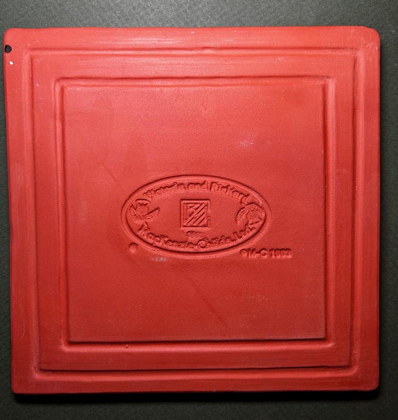 Może przedstawiać: Kwadratowa, czerwona płytka ceramiczna z zagłębioną obw&oacute;dką. Na środku owalny emblemat z napisem "Wom and Richard" i "Mackenzie-Childs Ltd.". Płytka oznaczona "PM-G 1992".