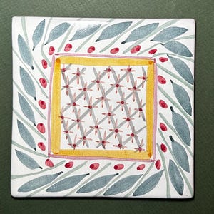 May include: Square ceramic tile with a hand-painted design. The tile features a central square with a grid pattern of gray flowers and red centers, framed by a yellow border. The outer edges have gray leaves and red berry-like accents.
