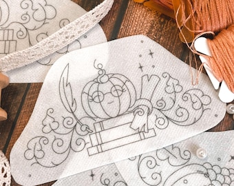May include: White fabric patches featuring black line drawings of pumpkins, books, and decorative swirls. The patches are on a wooden surface with orange thread spools, lace ribbon, and small white beads.
