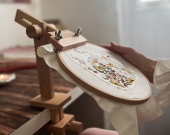 May include: A wooden embroidery hoop stand holds a round embroidery project with a floral design. The stand is clamped to a wooden table. The embroidery hoop is made of light-colored wood and holds a white fabric with a detailed floral pattern.