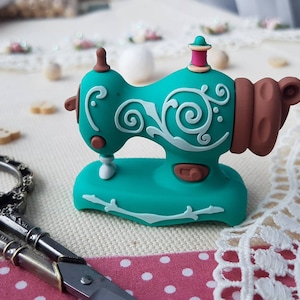 Può includere: Una figurina di macchina da cucire turchese con dettagli marroni e bianchi. La macchina da cucire in miniatura presenta decorazioni a volute e una bobina rosa e marrone. La figurina è posta su una superficie bianca con accessori per cucire.