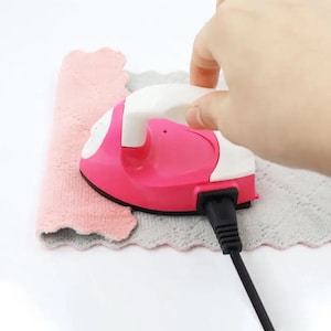 May include: A pink and white mini iron with a black power cord is shown on top of a pink and grey cloth. The iron is being held by a hand.