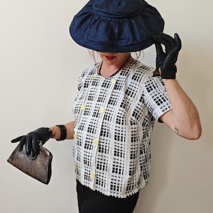 Puede incluir: Conjunto vintage con sombrero azul marino, guantes negros y blusa estampada. La blusa tiene un diseño de cuadrícula en blanco y negro con botones amarillos. Un bolso de mano marrón se sostiene en la mano izquierda. El atuendo se completa con pantalones negros.