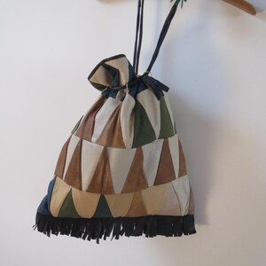 Puede incluir: Un bolso con cordón con un diseño de patchwork en tonos marrones, beige y verdes. El bolso tiene flecos negros en la parte inferior y cordones negros. Cuelga de una percha de madera.