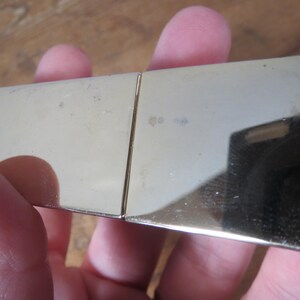 May include: A gold-colored, rectangular metal object with a hinged design. The surface reflects light, showing a mirror-like quality. The object appears to be a small, possibly decorative item.