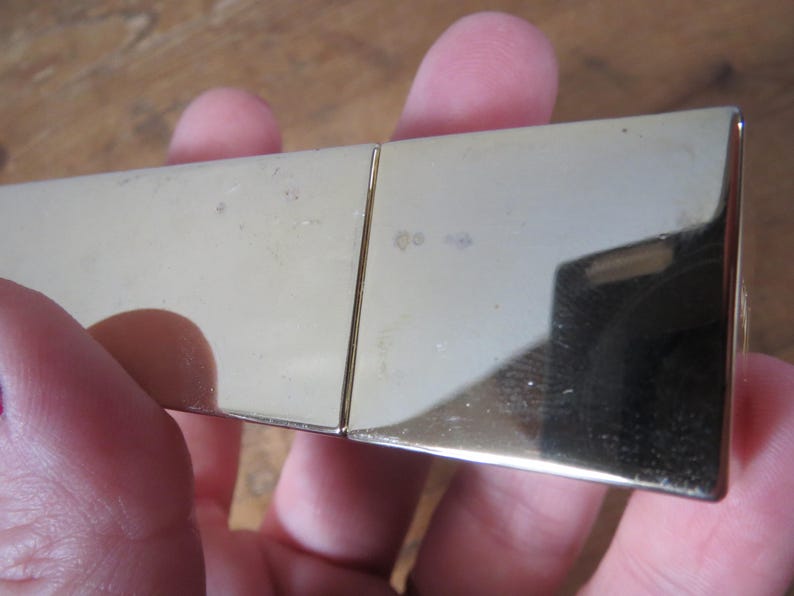 May include: A gold-colored, rectangular metal object with a hinged design. The surface reflects light, showing a mirror-like quality. The object appears to be a small, possibly decorative item.