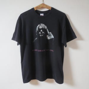 Puede incluir: Camiseta negra con una imagen en escala de grises de una persona haciendo una peineta. El texto "KURT COBAIN 1967-1994" está impreso en rosa debajo de la imagen. La camiseta está colgada en una percha de madera.