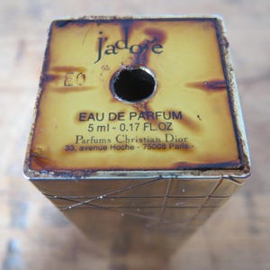 May include: A vintage perfume bottle with the text "J'adore" and "EAU DE PARFUM" printed on a yellow and brown surface. The bottle contains 5 ml - 0.17 FL.OZ of perfume and is from Parfums Christian Dior.