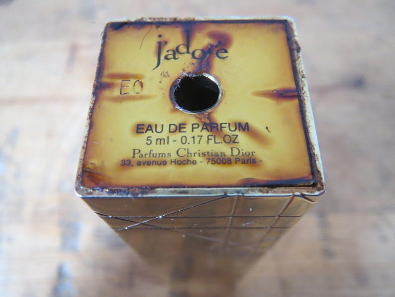 May include: A vintage perfume bottle with the text "J'adore" and "EAU DE PARFUM" printed on a yellow and brown surface. The bottle contains 5 ml - 0.17 FL.OZ of perfume and is from Parfums Christian Dior.