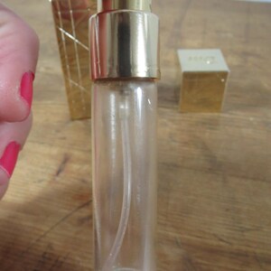 May include: A clear glass perfume bottle with a gold-colored cap and sprayer. The bottle is held by a person with red nail polish. Other gold-colored perfume bottles and a small box are in the background.