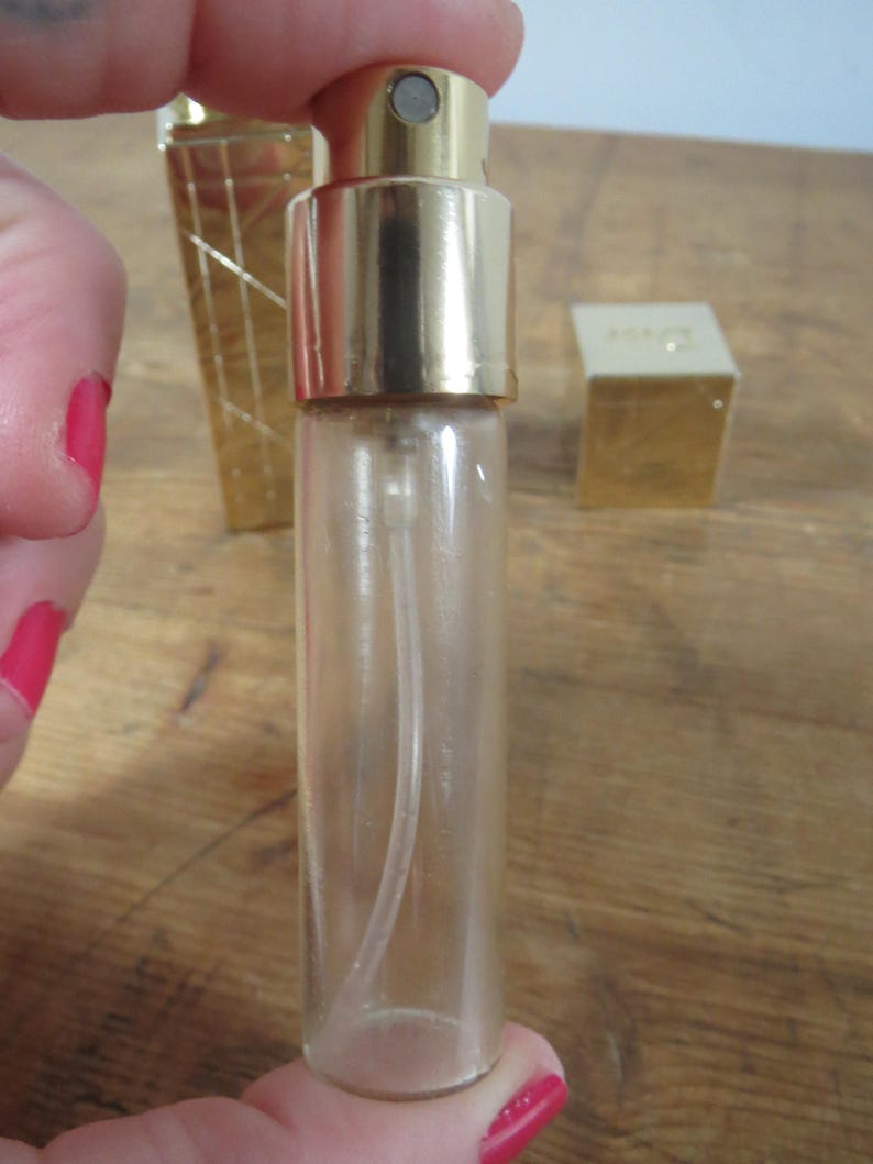 May include: A clear glass perfume bottle with a gold-colored cap and sprayer. The bottle is held by a person with red nail polish. Other gold-colored perfume bottles and a small box are in the background.