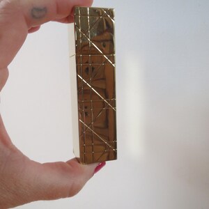 May include: A gold-colored rectangular lighter with a geometric pattern. The lighter is held in a hand against a white background. The lighter's surface has a textured, crisscross design.