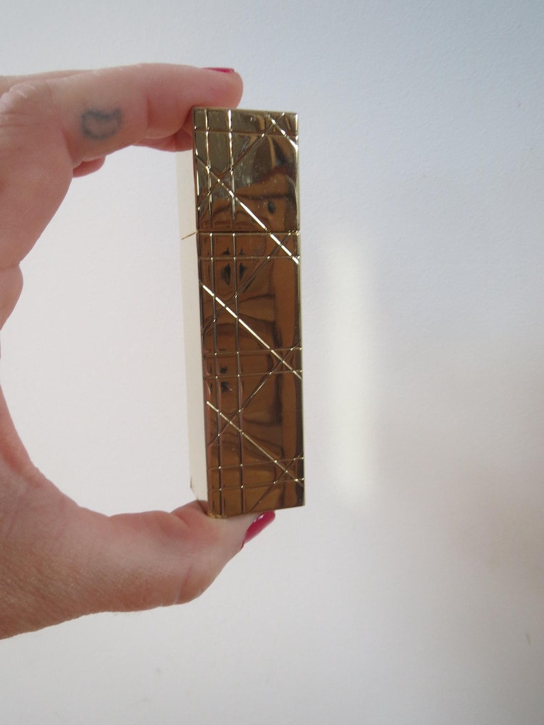 May include: A gold-colored rectangular lighter with a geometric pattern. The lighter is held in a hand against a white background. The lighter's surface has a textured, crisscross design.