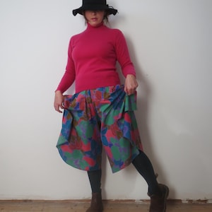May include: A person wearing a black hat, a pink turtleneck sweater, and colorful floral print culottes. The outfit is completed with dark leggings and brown boots. The culottes feature a mix of red, green, blue, and purple floral patterns.