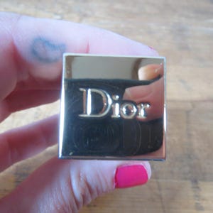 May include: A square, gold-toned compact with the word "Dior" in silver lettering. The compact is held in a hand with pink nail polish. The background is a light brown wooden surface.