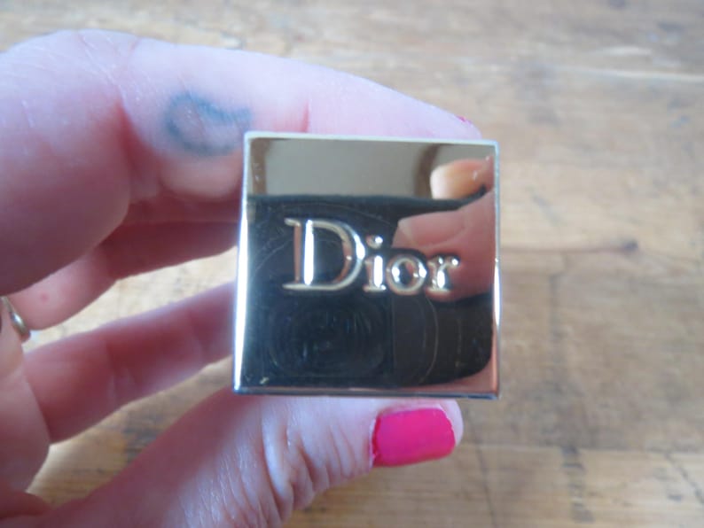 May include: A square, gold-toned compact with the word "Dior" in silver lettering. The compact is held in a hand with pink nail polish. The background is a light brown wooden surface.