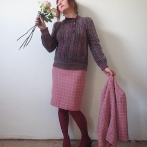 Può includere: Una donna indossa un maglione malva con collo abbottonato, una gonna rosa a quadri e calze bordeaux. Tiene in mano una giacca a quadri abbinata e un mazzo di rose rosa. Scarpe nere con dettagli dorati completano l'outfit. Stile vintage.