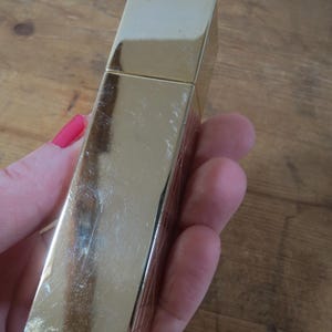 May include: A rectangular, gold-colored lighter with a hinged lid. The lighter has a shiny, reflective surface with some visible scratches. The lighter is held in a person's hand, with a wooden surface in the background.