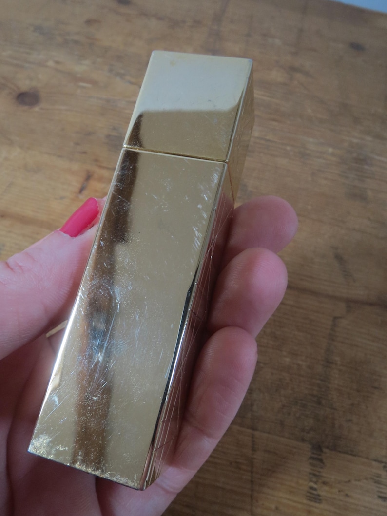 May include: A rectangular, gold-colored lighter with a hinged lid. The lighter has a shiny, reflective surface with some visible scratches. The lighter is held in a person's hand, with a wooden surface in the background.