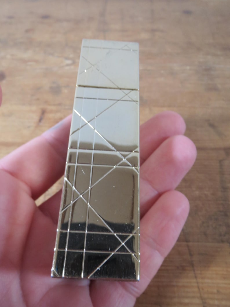 May include: A gold-colored rectangular lighter with a geometric pattern of intersecting lines. The lighter is held in a hand, and the background is a wooden surface.