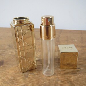 May include: A gold-colored perfume atomizer and case set. The atomizer is clear with a gold cap. The case is rectangular with a geometric design. A small gold box with the word "Dior" is also included.