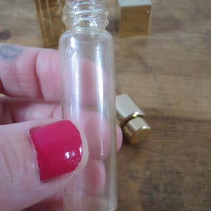May include: A clear, small glass perfume bottle with a threaded neck. The bottle is held in a hand with red nail polish. A gold-colored perfume bottle cap and a gold-colored box are in the background.